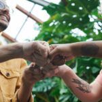 Four individuals clasping hands in a vibrant greenhouse filled with flourishing plants.